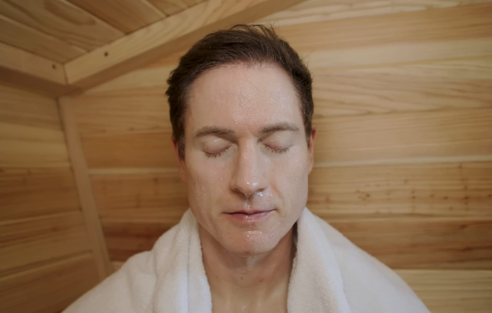 Bryan Johnson con los ojos cerrados y una toalla en los hombros dentro de su sauna de madera